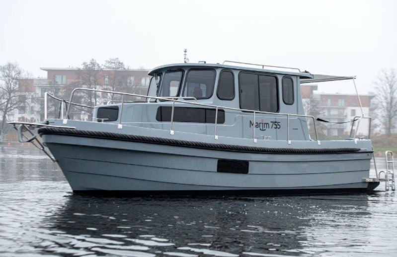 Ein graues Motorboot namens "Marim 755" liegt still in einem Hafen mit Gebäuden im Hintergrund.